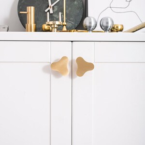 Op de afbeelding: Twee gouden kastknoppen met een uniek, driepuntig, bladvormig ontwerp. De knoppen zijn gemonteerd op een witte kastdeur.