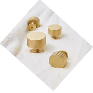 May include: Four gold-colored cabinet knobs with a cylindrical design and vertical grooved detailing. The knobs are set against a white, textured background, showcasing their metallic finish and simple elegance. These knobs are ideal for furniture upgrades.