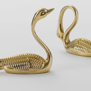 Könnte beinhalten: Zwei goldene Serviettenhalter in Schwanenform. Die Schwäne sind in entgegengesetzte Richtungen gerichtet und haben ein strukturiertes Design.