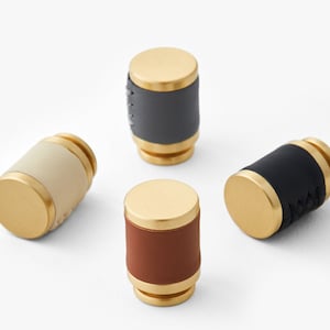 May include: Four cabinet knobs with a brass base and leather coverings in grey, beige, brown, and black.