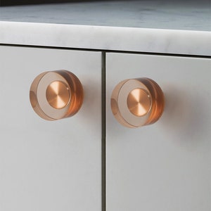 May include: Two round, clear, acrylic cabinet knobs with a copper coloured metal centre. The knobs are mounted on a white cabinet door.