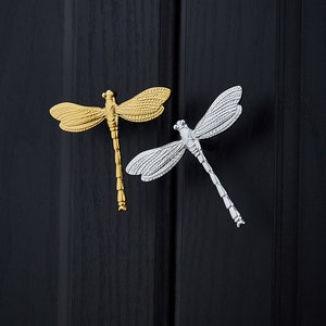 Könnte beinhalten: Zwei Schrankknöpfe in Libellenform, einer golden und einer silbern, vor schwarzem Hintergrund.