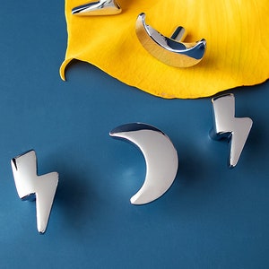 May include: Four silver cabinet knobs in the shapes of a crescent moon and a lightning bolt. The knobs are on a blue background.