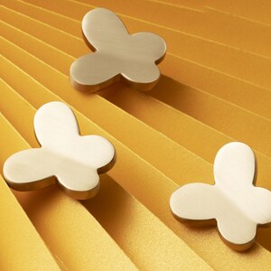 May include: Three butterfly-shaped cabinet knobs in a brushed brass finish. The knobs are arranged on a yellow, pleated background. The butterfly design features rounded edges and a simple, elegant aesthetic.