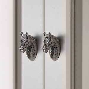 Pode incluir: Dois botões de porta de armário de cabeça de cavalo prateados. As cabeças de cavalo estão voltadas para a frente com a boca fechada. Os botões estão montados em uma porta branca.
