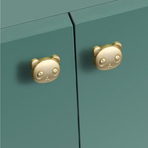 Puede incluir: Primer plano de un armario color turquesa con dos tiradores dorados en forma de panda. Los tiradores son redondos con un diseño de cara de panda, que incluye orejas, ojos y nariz. Las puertas del armario son de color turquesa liso.