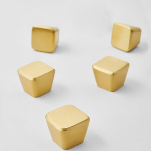 May include: Five gold cabinet knobs, each with a square top and a tapered body. The knobs are made of metal and have a matte finish. They are arranged on a white background.