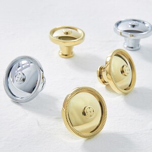 May include: Four cabinet knobs, two gold and two silver, with a decorative design in the center. The knobs have a raised edge and a textured surface.
