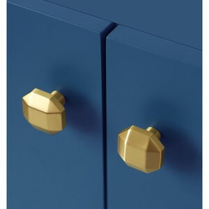 May include: Two gold-toned cabinet knobs with a geometric design, mounted on a blue cabinet door.