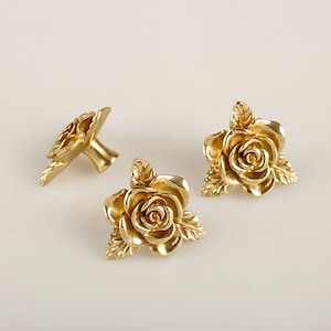 Puede incluir: Tres pomos de armario en forma de rosa de color dorado con detalles intrincados. Los pomos son de metal y tienen un acabado pulido.