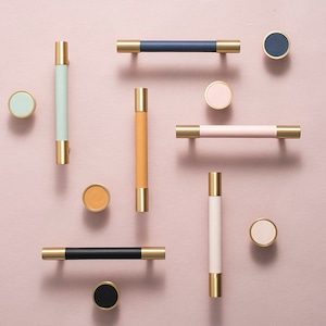 May include: Assortment of cabinet knobs and pulls in various colours, including black, white, pink, blue, green, and orange. The knobs and pulls have a gold finish and are arranged on a pink background.