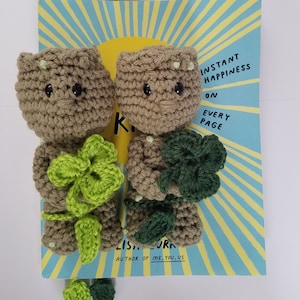 May include: Two crocheted brown and green characters holding green four-leaf clovers. The characters are standing on a book with the text "Instant Happiness on Every Page" and "Author of Me, You, Us".