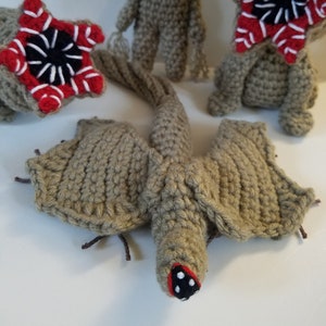 May include: Three crocheted amigurumi toys inspired by the Demogorgon from the show Stranger Things. The toys are beige with red and black details. One toy is a Demogorgon with a long tail and wings.