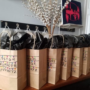 May include: Eight brown paper gift bags with black tissue paper inside. Each bag has the alphabet written on it with colourful lights, like Christmas lights, above the letters. The letters are in order, with each bag containing a portion of the alphabet. The bags are on a wooden table.