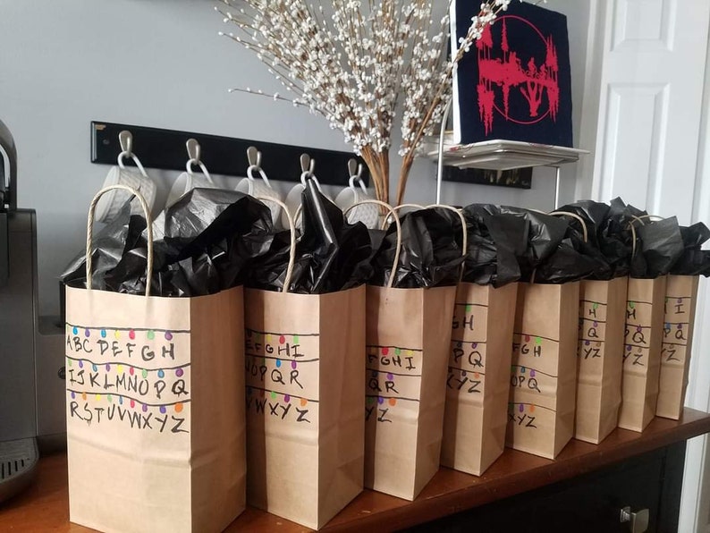 May include: Eight brown paper gift bags with black tissue paper inside. Each bag has the alphabet written on it with colourful lights, like Christmas lights, above the letters. The letters are in order, with each bag containing a portion of the alphabet. The bags are on a wooden table.