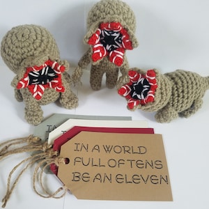 May include: Three crocheted plush toys shaped like the Demogorgon from the show Stranger Things. The toys are beige with red and black details. The toys are sitting on a brown surface with a tag that reads "In a world full of tens be an eleven".