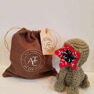 May include: A green crocheted plush toy with a red and black mouth, resembling a monster from the show Stranger Things. The toy is sitting on a white surface next to a brown drawstring bag with a white logo that reads "All Tangled Creations Co."