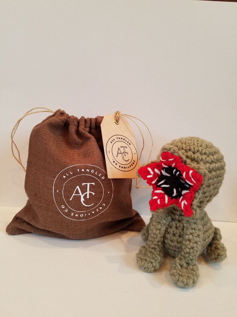 May include: A green crocheted plush toy with a red and black mouth, resembling a monster from the show Stranger Things. The toy is sitting on a white surface next to a brown drawstring bag with a white logo that reads "All Tangled Creations Co."