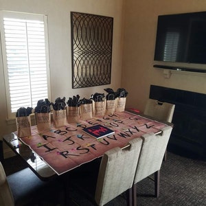 May include: A brown wooden table with a game board featuring the alphabet and a red figure. There are brown paper bags with black handles on the table. The bags are arranged in a row. The table is in a room with a fireplace and a flat screen TV.