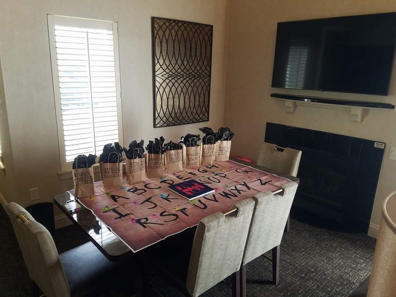 May include: A brown wooden table with a game board featuring the alphabet and a red figure. There are brown paper bags with black handles on the table. The bags are arranged in a row. The table is in a room with a fireplace and a flat screen TV.