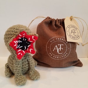 May include: A crocheted green and red monster plush toy with a large mouth and teeth. The toy is sitting on a white surface next to a brown burlap bag with a white logo that says "All Tangled Creations" and a brown tag with the same logo.