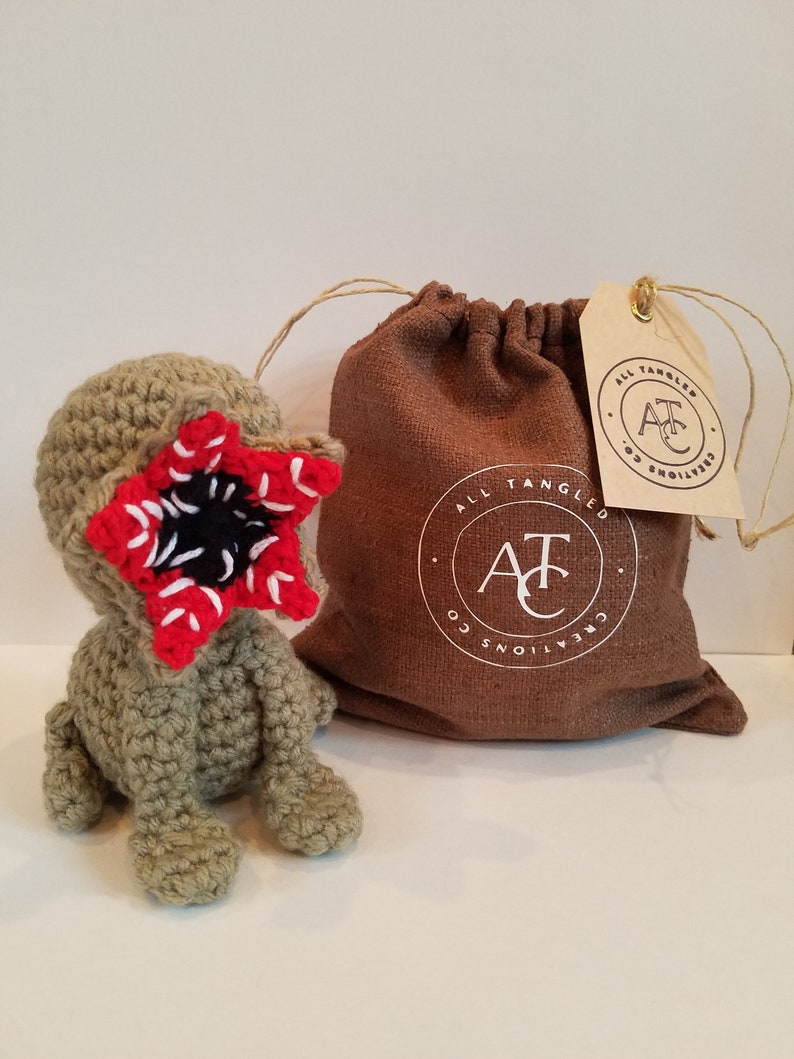 May include: A crocheted green and red monster plush toy with a large mouth and teeth. The toy is sitting on a white surface next to a brown burlap bag with a white logo that says "All Tangled Creations" and a brown tag with the same logo.