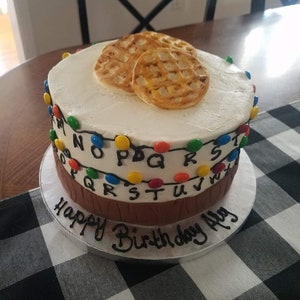May include: A two-tiered white cake decorated with a brown tree trunk base, colourful lights, and the letters of the alphabet. The top tier has two Eggo waffles on top. The cake is on a silver cake stand and has the words "Happy Birthday Ally" written in black icing.