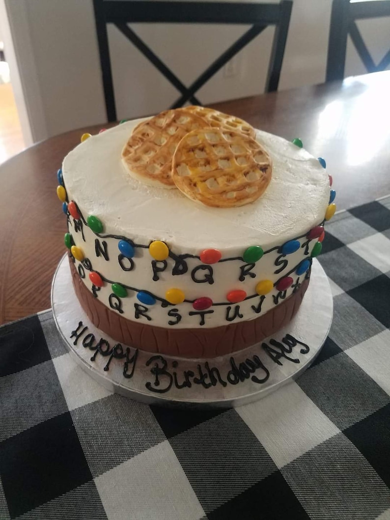 May include: A two-tiered white cake decorated with a brown tree trunk base, colourful lights, and the letters of the alphabet. The top tier has two Eggo waffles on top. The cake is on a silver cake stand and has the words "Happy Birthday Ally" written in black icing.
