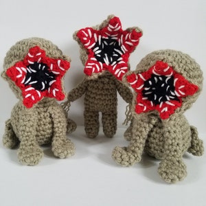 May include: Three crocheted plush toys shaped like the Demogorgon from the show Stranger Things. Each toy has a red and white mouth with black teeth and a beige body.