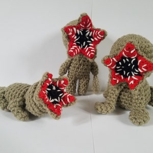 May include: Three crocheted plush toys shaped like the Demogorgon from the show Stranger Things. The toys are beige with red and white details. The Demogorgon's mouth is open and has black teeth.