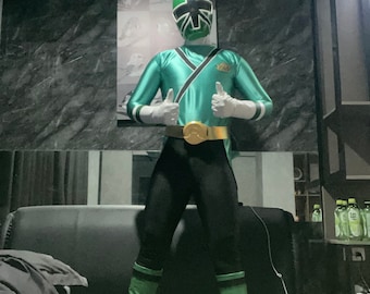 Puede incluir: Una persona vistiendo un traje de Power Ranger verde y negro con un cinturón dorado y botas verdes. El traje tiene un símbolo blanco y negro en el pecho.