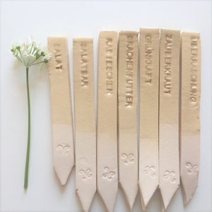 Könnte beinhalten: Acht beigefarbene Pflanzenmarkierer mit spitz zulaufenden Enden und einem dekorativen Design am unteren Ende. Die Markierer sind mit folgendem Text beschriftet: "SALAT", "SALATEAR", "RADIESCHEN", "DRACHENFUTTER", "GRÜNKRAFT", "ZAUBERKRAUT", "LIEBE & SORGEN".