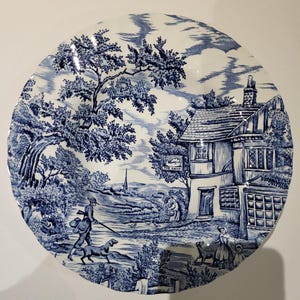 May include: A blue and white decorative plate with a pastoral scene. The plate features a detailed illustration of a man with a dog, a building with the text "The Swan", and trees. The plate has a classic, vintage aesthetic.