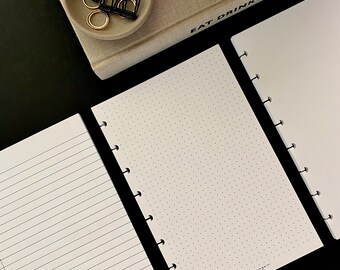 Op de afbeelding: Drie verschillende notitieboekjes met witte pagina's en zwarte omslagen. De notitieboekjes hebben verschillende patronen op de omslagen: een heeft lijnen, een heeft stippen en een is leeg. De notitieboekjes liggen op een zwart oppervlak. De tekst op de omslagen luidt "noted" en "dotted".