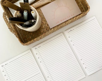 May include: Three white lined pages with holes on the left side. The pages are labeled "to do" and "notes". The pages are on a white surface. A wicker tray with various items is in the background.