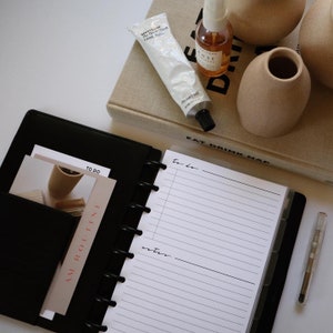 May include: A black leather planner with a white page open to a to-do list and notes section. The planner has a pocket on the left side with a card that says "AM. ROLLINI". There is a clear pen on the right side of the planner.
