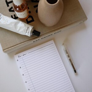 May include: A white lined notepad with the words "to do" written on it. A clear pen with a silver tip is lying on a white surface next to the notepad. A book with the words "Eat Drink Nap" is visible in the background.