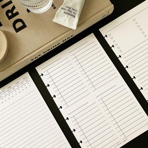 Op de afbeelding: Een witte planner met een zwart-witte kalender op de linkerpagina en een gelinieerde pagina aan de rechterkant. De planner heeft een zwarte binding met zilveren ringen. De tekst "EAT DRINK NAP" staat gedrukt op de kaft van de planner.
