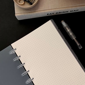 May include: A white, dotted notebook with a clear plastic binding and a gray cover. The notebook is open to a blank page. The text "EAT DRINK NAP" is visible on a beige book in the background.