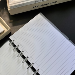 May include: A white lined notebook with a black and clear plastic binding. The notebook has a clear plastic cover with tabs on the side. The notebook is lying on a black surface.