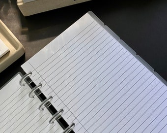 Op de afbeelding: Een wit, gestreept notitieboek met een zwarte en doorzichtige plastic binding. Het notitieboek heeft een doorzichtige plastic omslag met tabs aan de zijkant. Het notitieboek ligt op een zwart oppervlak.