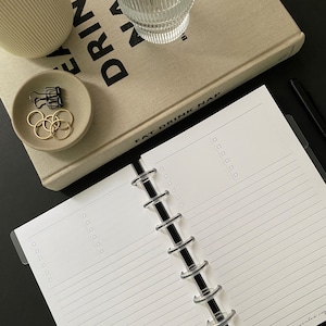May include: An open, white, lined notebook with a black ring binder. The notebook is open to a blank page with lines. The notebook is on a black surface. The text "EAT DRINK NAP" is visible on the cover of a book in the background.