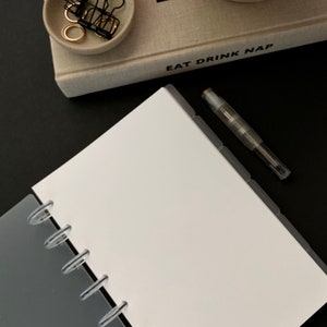 May include: A white notebook with a gray cover and clear plastic binding rings. The notebook has a black pen and a tabbed divider. The text "eat drink nap" is visible on the cover of a book in the background.