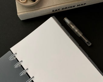 May include: A white notebook with a gray cover and clear plastic binding rings. The notebook has a black pen and a tabbed divider. The text "eat drink nap" is visible on the cover of a book in the background.