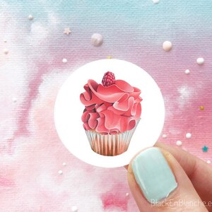 Puede incluir: Un cupcake rosa con una frambuesa en la parte superior. El cupcake está decorado con glaseado rosa y tiene un forro de papel blanco. El cupcake está sobre un fondo blanco.