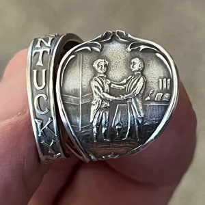 Op de afbeelding: Zilveren ring met een gedetailleerde gravure van twee mannen die elkaar de hand schudden. De ring is aan de zijkant gegraveerd met het woord "TUCK".