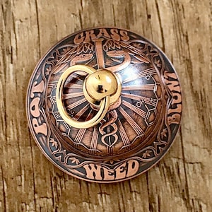 Può includere: Un oggetto rotondo in metallo color rame con un anello e una sfera dorati. L'oggetto presenta le parole "PAR" e "WEED", e "GRASS BLUNT". Il design include un simbolo medico e un motivo a raggi solari.