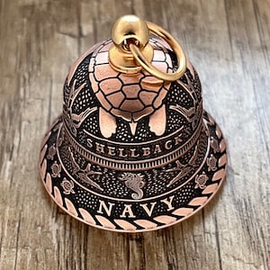 Peut inclure: Une cloche de couleur cuivre avec un sommet doré et un anneau. La cloche est décorée d'une tortue de mer, d'hippocampes et des mots "Shellback Navy".