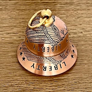 May include: A copper-colored bell charm featuring an engraved design of a person wearing a feathered headdress. The word LIBERTY is etched twice around the bell, accompanied by small black stars. A gold-toned metal jump ring is attached to the top.