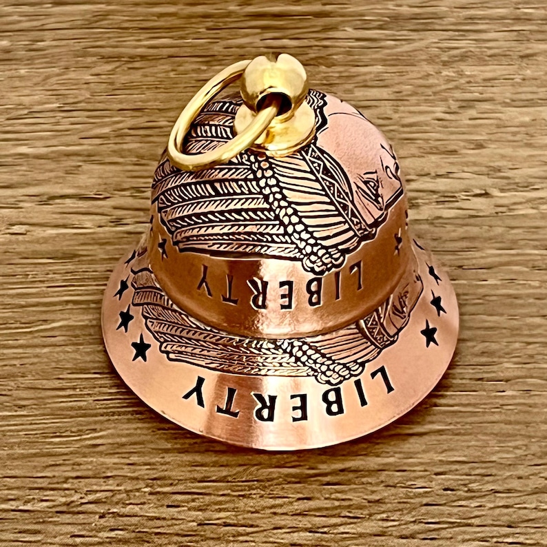 May include: A copper-colored bell charm featuring an engraved design of a person wearing a feathered headdress. The word LIBERTY is etched twice around the bell, accompanied by small black stars. A gold-toned metal jump ring is attached to the top.